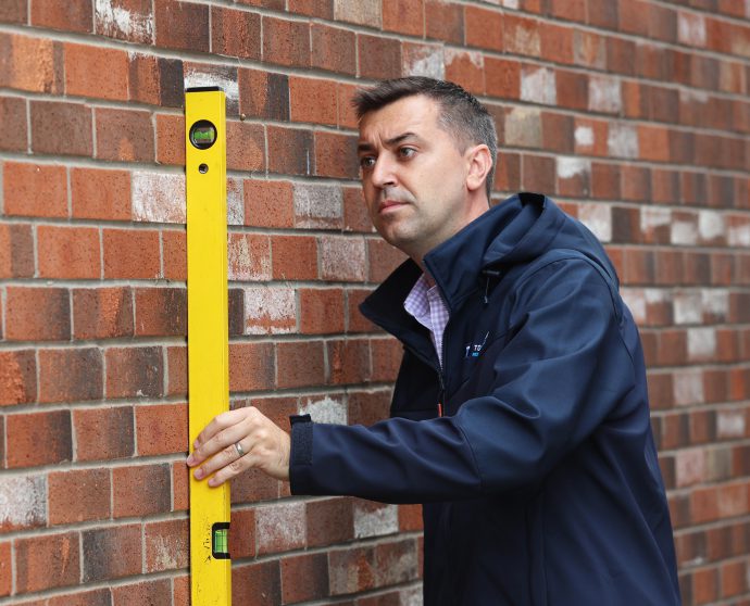 A man holds a yellow level against a brick wall to ensure it is straight.